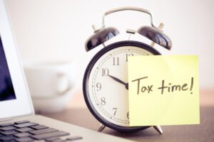 a clock with a post-it note that says "Tax time!" next to a laptop computer.